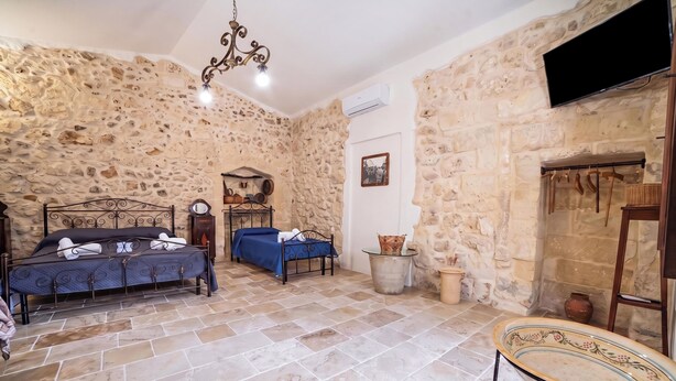 Habitación con paredes de piedra, dos camas con estructura metálica, una lámpara colgante, una mesita y una chimenea decorada con objetos colgantes.