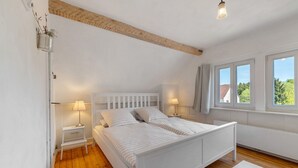 Ett sovrum med en vit säng, två sänglampor, ett trägolv och ett fönster med utsikt över grönområden.
