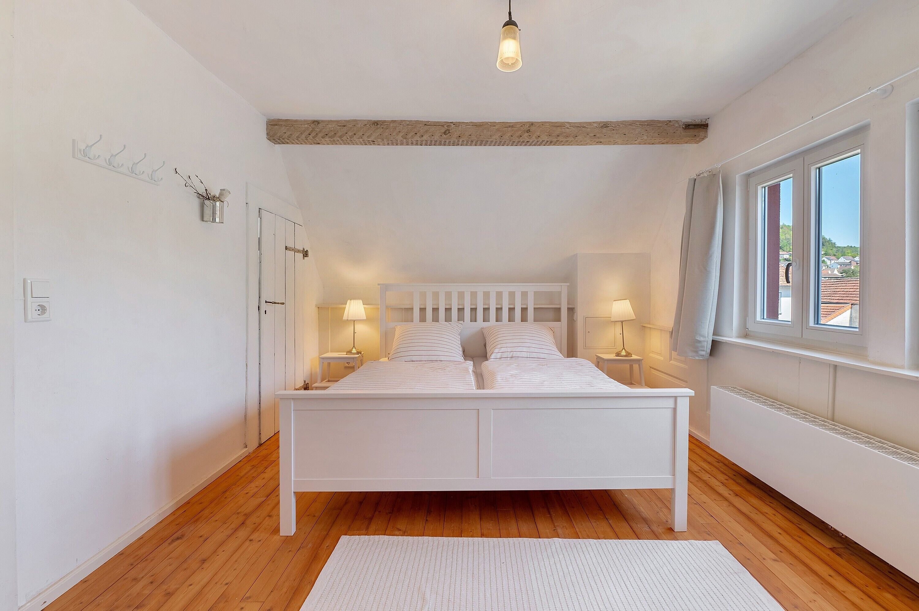 Een slaapkamer met een wit bed, houten vloer, een hanglamp en een raam met gordijnen.