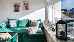 A green sofa with decorative pillows, a glass table with a candle, and a lantern on a balcony with a view of houses.