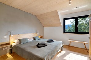 Une chambre avec un plafond en pente, un lit avec une tête de lit, des tables de chevet, un banc et une fenêtre donnant sur un beau paysage.