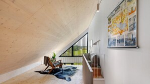 Un espacioso dormitorio tipo loft con techo inclinado, un ventanal grande, piso de madera, alfombra blanco y negro, una silla de madera y un mapa colgado en la pared.