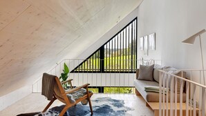 Un dormitorio tipo loft moderno con techo inclinado, una silla de madera, un sofá, un ventanal y vistas a zonas verdes.
