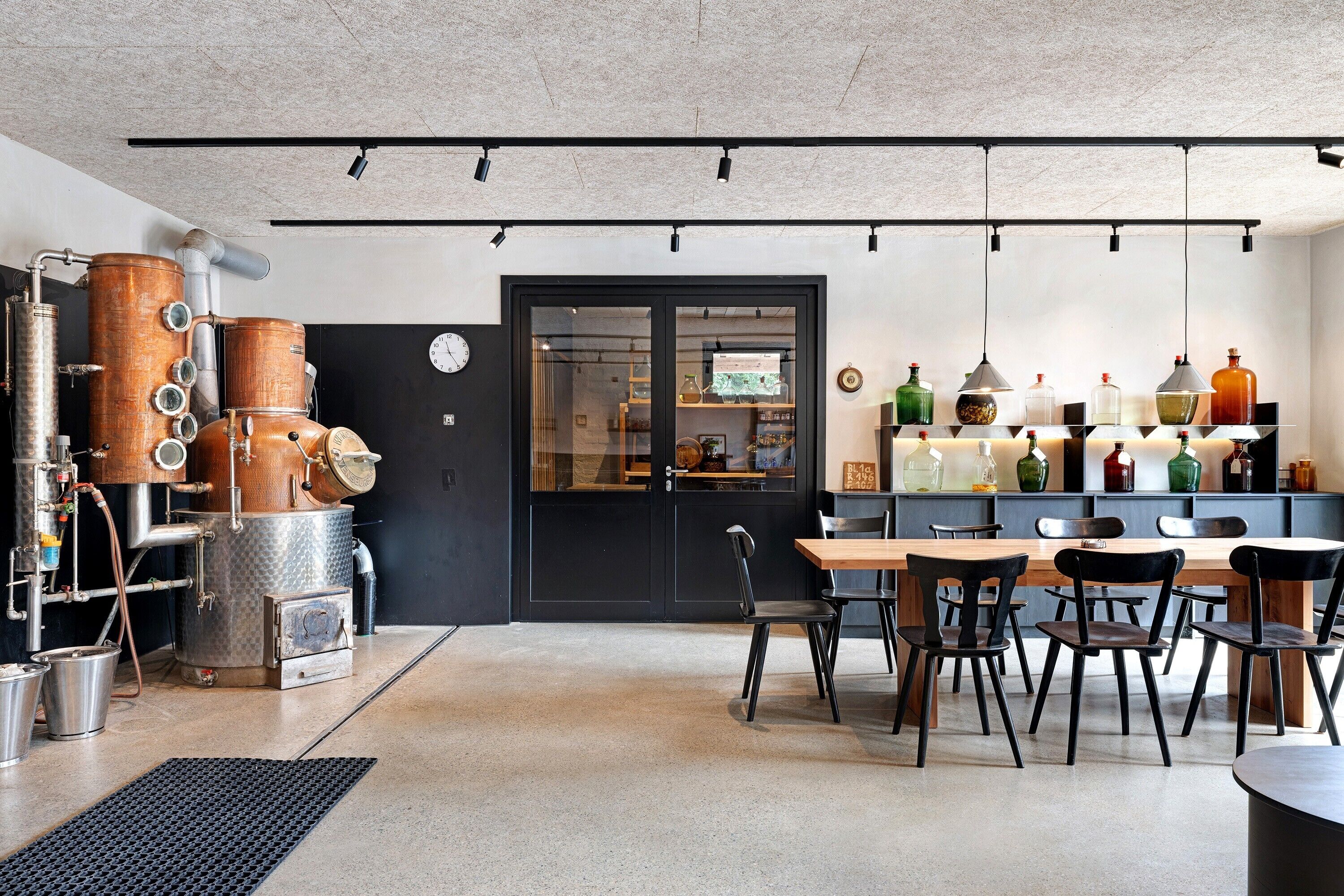 A modern kitchen with industrial equipment, a dining area with chairs and a table, and a display of colorful bottles on shelves.