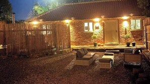 A rustic stone building with a wooden door, a small porch with seating, and a wooden fence.