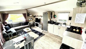 Interior compacto de una autocaravana con sofá, mesa de comedor, zona de cocina y una pequeña ventana con cortinas.