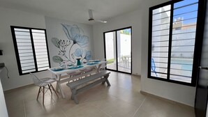 Uma área de refeições com uma mesa posta para quatro pessoas, um banco e uma cadeira. Há grandes janelas com persianas, um ventilador de teto e vista para o exterior.