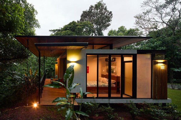 A modern, single-story house with a wooden deck, a bedroom with a bed and nightstand, and a bathroom with a shower.