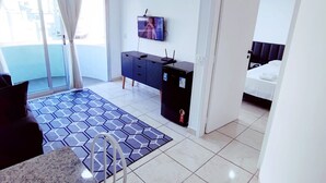 A modern hotel room with a large window, a flat-screen TV, a dark-colored dresser, a bed with a white headboard, and a small kitchen area with a granite countertop and a chair. - APARTAMENTO O PREFERIDO LIBERDADE SP (São Paulo)