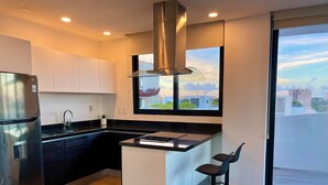 Une cuisine moderne dotée d’un réfrigérateur en acier inoxydable, de plans de travail noirs, d’armoires blanches, d’une grande fenêtre offrant une vue sur la ville et de deux tabourets de bar.