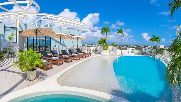Un área de piscina moderna con sillones de playa y sombrillas, rodeada de palmeras y un cielo azul despejado.
