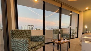 Habitación de hotel moderna con balcón, dos sillones estampados, una pequeña mesa de madera con una botella y un vaso, y un ventanal con vistas a la ciudad.