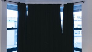 Habitación de esquina con una mesa y banco de madera, ventanas amplias con cortinas negras, y vista a edificios.