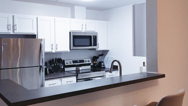 Una cocina moderna con armarios blancos, un refrigerador de acero inoxidable, un microondas, un horno y un lavamanos. Hay dos taburetes de bar en la encimera.