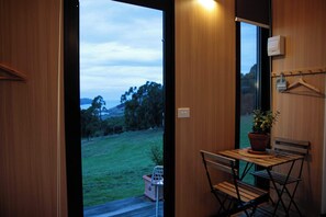 Una habitación con puerta de madera, una mesa pequeña con una silla, una planta colgante y vistas a un espacio cubierto de césped.