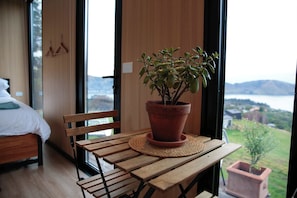 A room with a bed, a wooden bench, a potted plant on a woven mat, and a view of a body of water and hills. - Bayview Paradise by Tiny Away (Diamond Harbour)