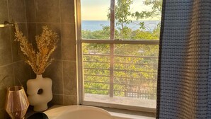 A bathroom with a bathtub, a window with a view of greenery and a body of water, and a vase with dried plants. - house with a beautiful sea view in enchanting SurfCity with WiFi, AC and Pool. (La Libertad)