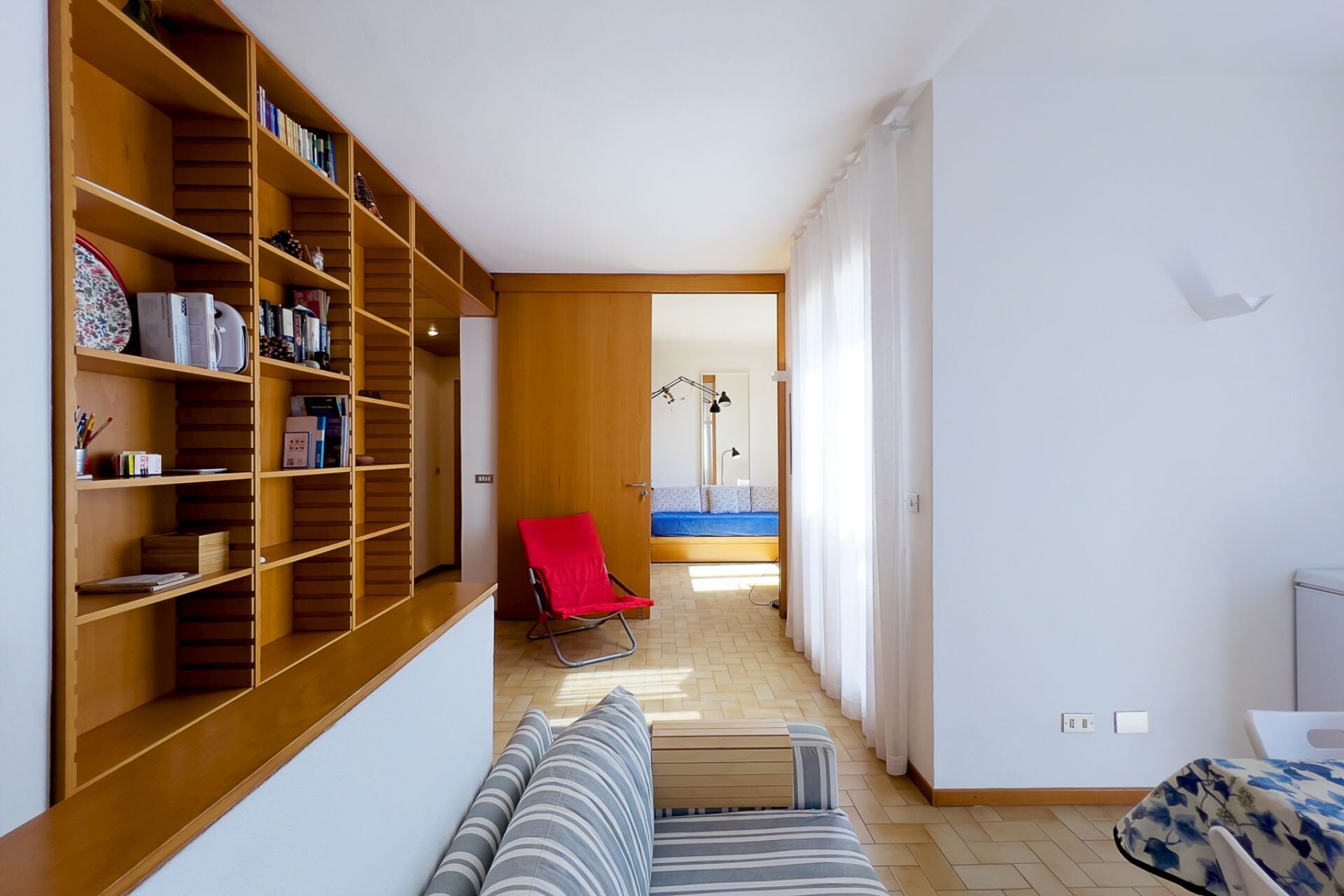 A narrow hallway with a wooden bookshelf, a red chair, a sofa, and a bed with a blue mattress.