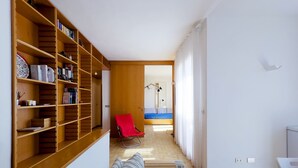 A narrow hallway with a wooden bookshelf, a red chair, a sofa, and a bed with a blue mattress. - CASA LUISA A CAPO'DARCO - CASA LUISA (Rio Marina)