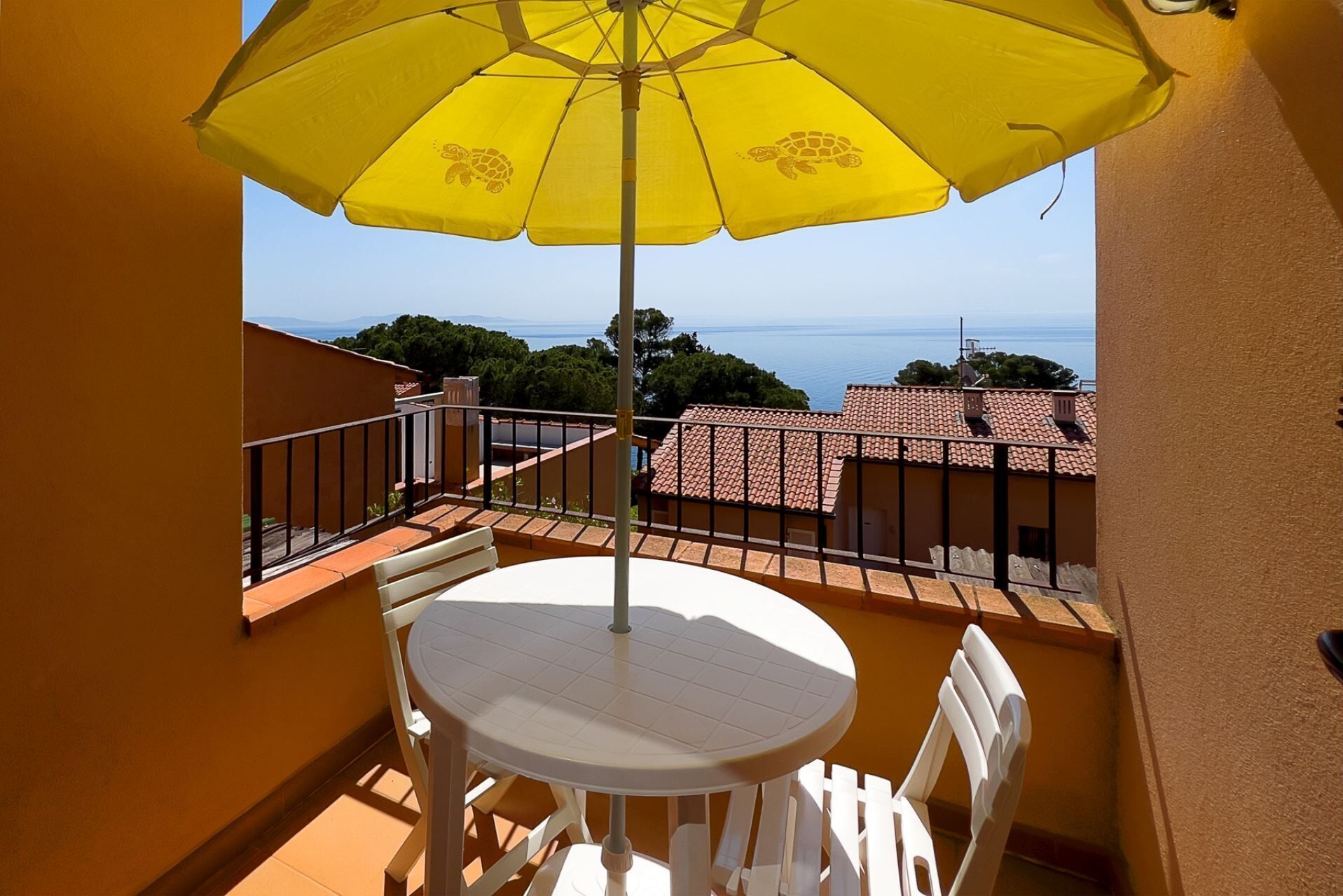 A balcony with a yellow umbrella, white table, and chairs, overlooking a coastal area with buildings and trees.