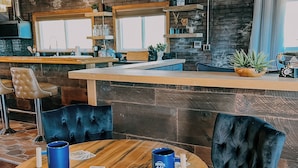 Un rincón acogedor con una barra de madera, taburetes con cojines y una mesa redonda de madera con una taza azul y cartas.