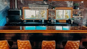 Una zona de bar con mostrador de madera, taburetes de cuero, pared de ladrillo y un ventanal grande.
