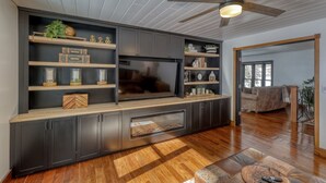 A modern living room with a large flat-screen TV, wooden shelves, and a ceiling fan. - Calamity Jane’s Hidden Haven – 12.5 private acres, multiple patios, fire pit & 4 king suites! (Lead)
