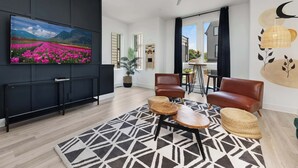 A modern living room with a large flat-screen TV, black and white patterned rug, two leather chairs, and a ceiling fan.