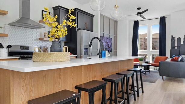 A modern kitchen with a central island, bar stools, pendant lighting, and a dining area with a sofa and chairs.