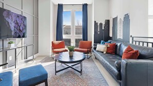 A modern living room with a black leather sofa, two red armchairs, a round coffee table, and a flat-screen TV mounted on the wall.