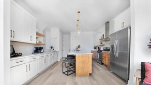A modern kitchen with white cabinets, a central island, bar stools, a stainless steel refrigerator, and a pendant light. - Camellia | Rooftop & Pet-Friendly Stay (Nashville)