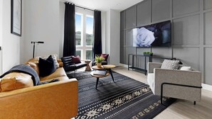 A modern living room with a sofa, armchair, and coffee table, a flat-screen TV mounted on the wall, and a large window with curtains. - The Bluestem - Stylish Design, Rooftop w/ Firepit! (Nashville)