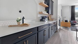 A modern kitchen with black cabinets, a white countertop, and a wooden cutting board. There is a potted plant on the counter and a coffee machine. - The Bluestem - Stylish Design, Rooftop w/ Firepit! (Nashville)
