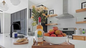 A modern kitchen with a white countertop, a bottle of Wildthings wine, a bowl of fruit, and a decorative vase.