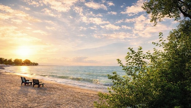 Ein Strand mit zwei Holzstühlen und einem Tisch, ein sandiger Uferstreifen und Blick auf Meer und Himmel.