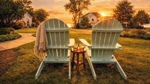 Zwei Adirondack-Stühle mit einem Tisch dazwischen, auf einer grünen Wiese mit Sonnenuntergang im Hintergrund.