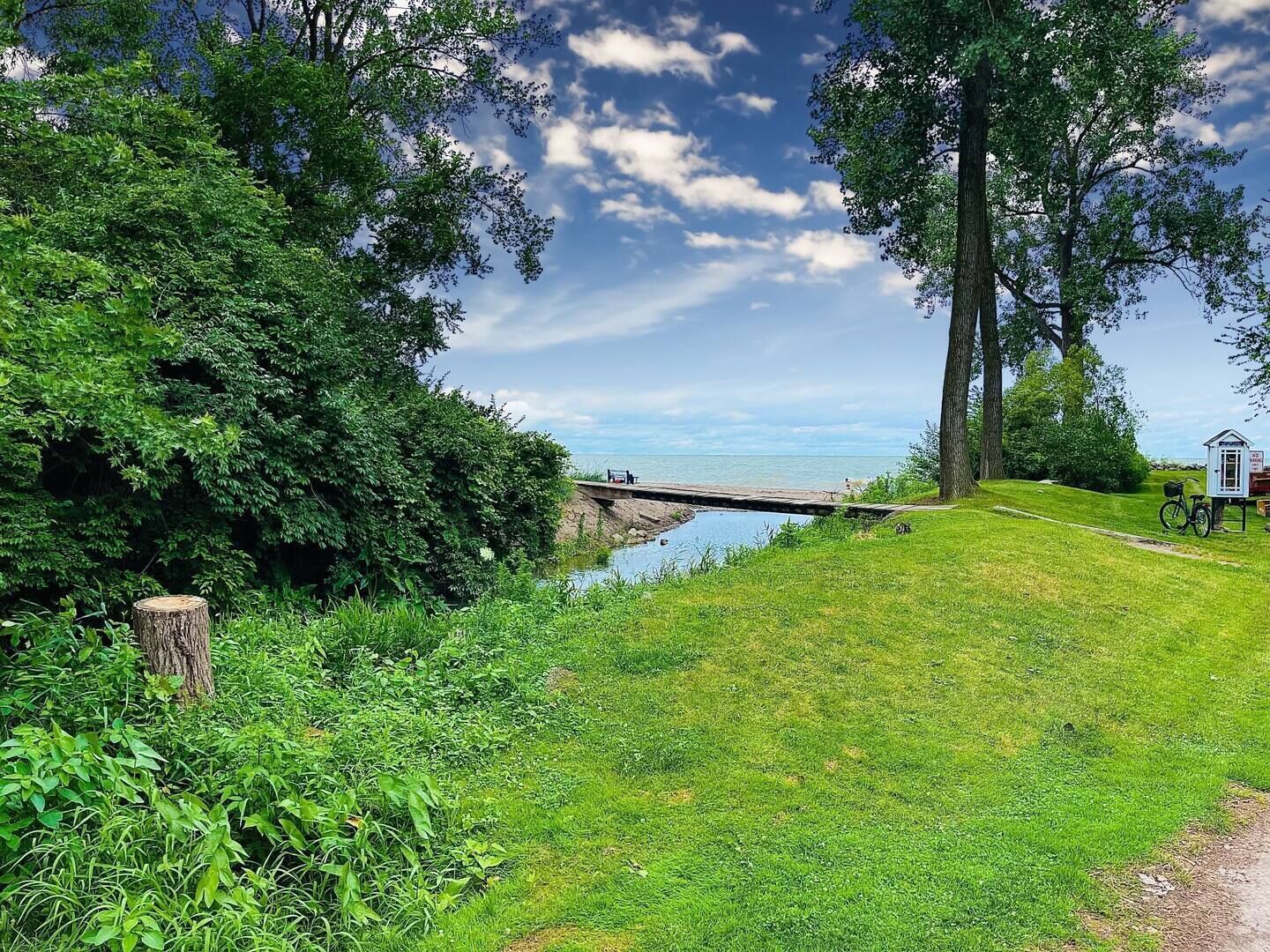 A grassy area with a small wooden bridge over a body of water, a bicycle, and a small white structure.