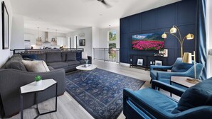 Un salon moderne avec un canapé, une table basse et des fauteuils bleus qui apportent une touche de couleur. Un téléviseur est fixé au mur, et il y a un balcon avec une balustrade.