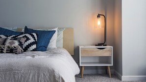 A neatly made bed with a white comforter and patterned pillows, a bedside table with a lamp, and a light-colored wall.