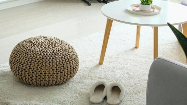 A modern living room with a TV, a guitar, a round table with a plant, a woven pouf, and slippers on a rug.