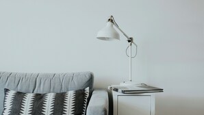 A grey sofa with patterned cushions, a white side table with a lamp, and a dark wooden floor.
