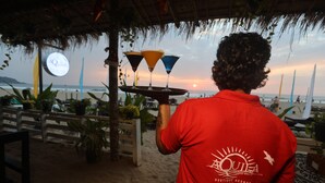 Une personne vêtue d’une chemise rouge tient un plateau avec trois boissons dans un endroit situé au bord de la plage, doté d’un toit de chaume et offrant une vue sur l’océan.