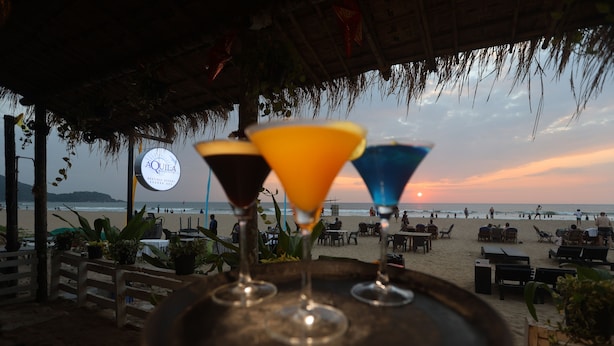 Un bar situé en bord de mer, avec un toit de chaume, une enseigne indiquant « AQUILA » et trois verres à martini colorés disposés sur un plateau.