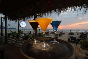 Un bar frente a la playa con techo de paja, un letrero que dice "AQUILA" y tres coloridos vasos de martini en una bandeja.