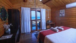 Une chambre rustique en bois, avec un lit, un bureau, une chaise et une vue sur l’océan par une porte vitrée.
