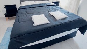 Ein modernes Schlafzimmer mit einem großen Bett, einem grauen Kopfteil, weißen Kissen und einem blauen Zierkissen.