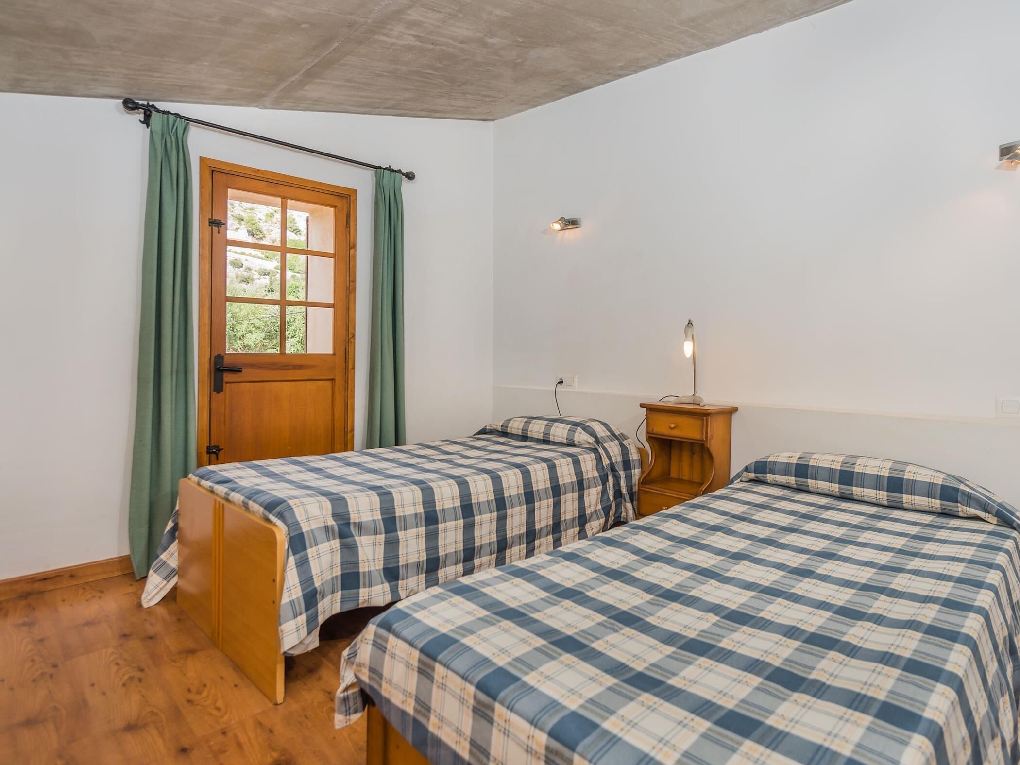 Una habitación con dos camas, una puerta de madera, una mesita de noche pequeña con una lámpara y una ventana con cortinas.