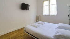 Un lit simple avec du linge de lit blanc, un téléviseur fixé au mur, une fenêtre avec des rideaux légers et un radiateur.