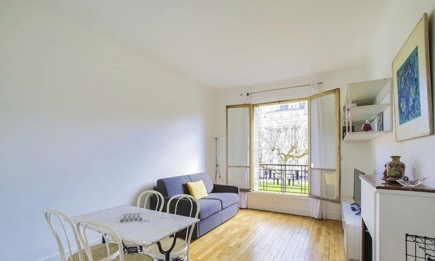 A room with a dining table, chairs, a sofa, a window with a view of trees, and a small kitchen area.