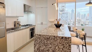 Una cocina moderna con armarios blancos, encimera de granito, estufa a gas y vista a un paisaje urbano a través de un ventanal amplio.